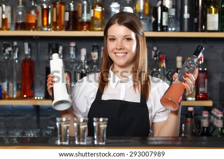 Portrait of a pretty bartender standing smiling and holding two bottles of liquors, shelves full of bottles with alcohol on the background