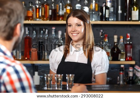 Portrait of a pretty bartender standing smiling and looking at her visitor sitting back to us at the counter talking to her, shelves full of bottles with alcohol on the background
