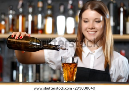 Portrait of a pretty bartender standing smiling and pouring a drink from a bottle, shelves full of bottles with alcohol on the background