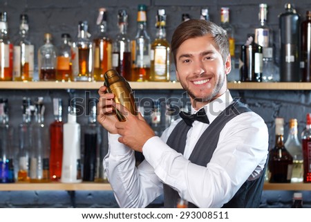 Portrait of a handsome bartender standing at the counter smiling holding a golden shaker and mixing a drink, shelves full of bottles with alcohol on the background