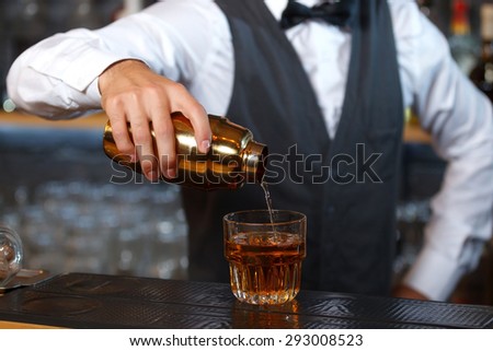 Close up photo of a bartender holding a golden shaker in his hand and pouring a cocktail in a low wide glass, shelves full of bottles with alcohol on the background