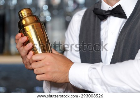 Close up photo of a bartender standing at the counter holding a golden shaker and mixing a drink, shelves full of bottles with alcohol on the background