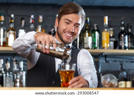 Portrait of a handsome bartender standing at the counter smiling and pouring a drink from a shaker to a glass, shelves full of bottles with alcohol on the background