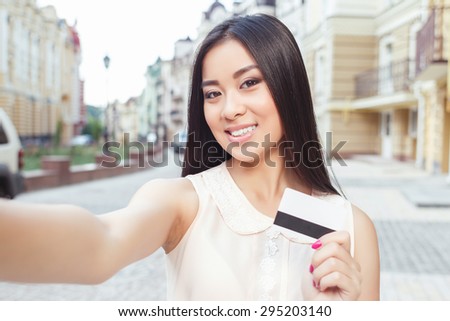 Portrait of a beautiful asian girl with long hair, wearing beige blouse standing smiling and holding a credit card in her hand and making a selfie