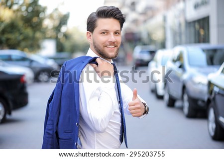 Be positive. Vivacious handsome young bearded businessman holding his jacket on the shoulder and smiling while thumbing up.