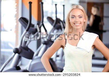 Waist up portrait of a beautiful young blond girl wearing a white top standing smiling in a fitness club holding her hands on her hips, with a towel on her shoulder