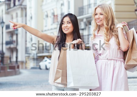 Friends on shopping. Portrait of asiang girl and her blond friend standing on the street holding shopping bags and pointing somewhere , beautiful old city on the background