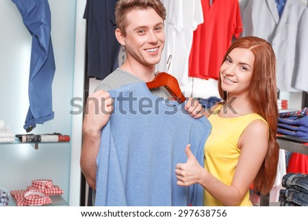 Shopaholic. Smiling customer holding hanger with colorful clothe in retail store and saleswoman thumbing up