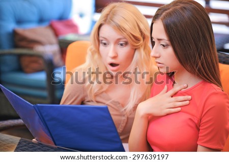 Shocking prices. Two pretty girlfriends sitting in cafe and  shocked from looking through price list