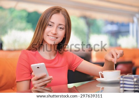 Pleasant time. Yong pretty woman sitting in cafe mixing sugar in cup with coffee and holding  mobile phone