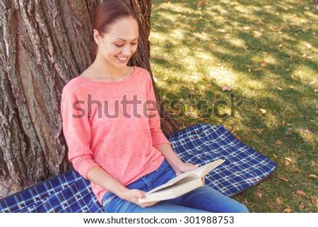 Funny story. Young pretty smiling woman sitting on cover at tree in park and reading interesting book.