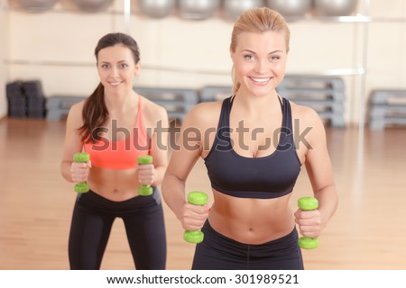 Holding hard. Two persistent women doing weights fitness exercises by using little dumbbells in gym