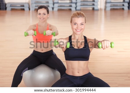 In great shape. Pair of women sitting on fitness balls in gym and doing exercises with little dumbbells.