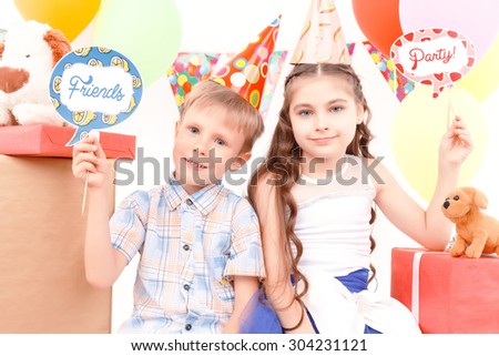 Birthday kids. Cheerful boy and girl sitting on background of presents holding tags with birthday related signs during party.