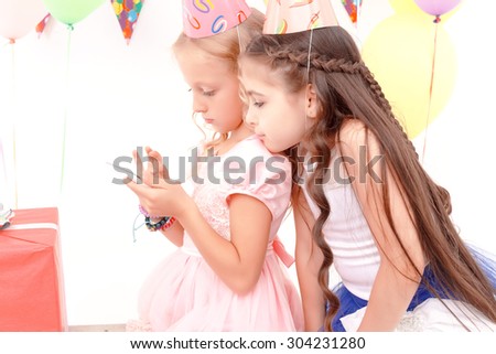 Exciting game. One little pretty girl holding mobile phone and another looking at it during birthday party.