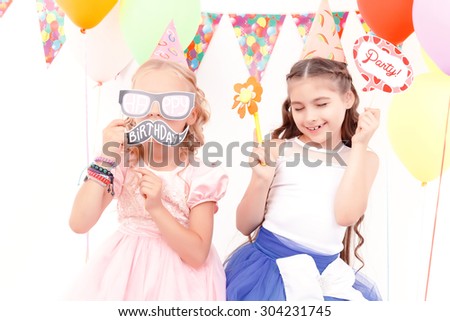 In full play. Portrait of two little joyful girls, one of them covering her face with birthday tag and paper glasses.