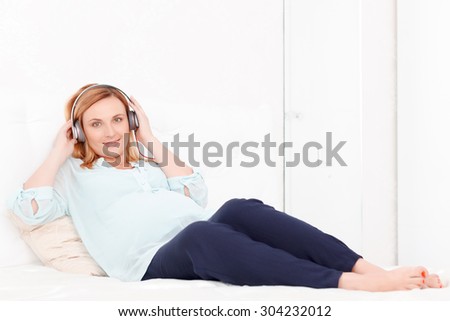 Relaxing moment. Portrait of pretty blond-haired pregnant woman lying on bed and listening to music.