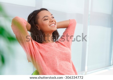 Pleasant moment. Portrait of youthful attractive mulatto woman sitting in office with closed eyes and arms behind her head.