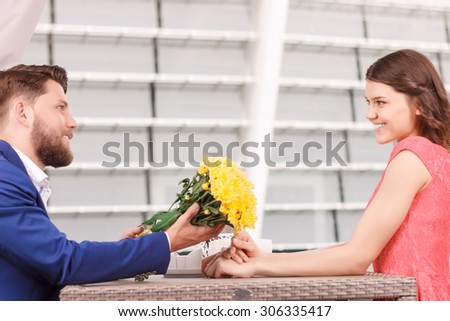 To you. Handsome young man with beard giving beautiful flowers to his pretty smiling girlfriend in restaurant.