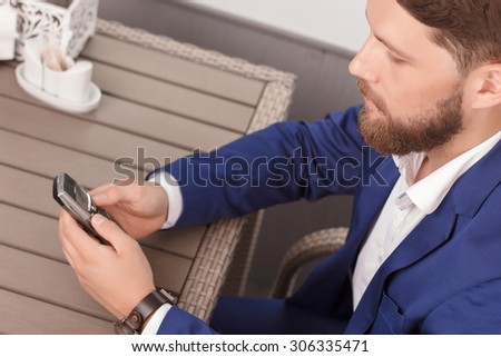 Checking messages. Portrait of young-looking handsome man with beard sitting in restaurant and using his mobile phone