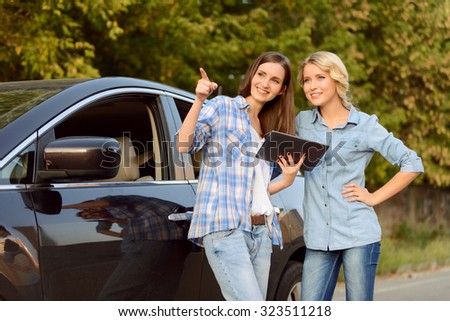 Look here. Cheerful young contented girls holding laptop and looking up while standing near the car