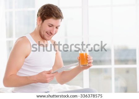Day planning. Young good-looking man sits on bed and checks his day-planner while drinking fresh fruit juice.