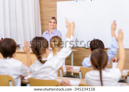In the school. Young female teacher is looking at her pupils while they all raising their hands in the air.