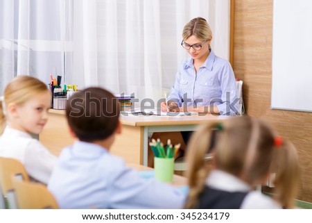 In the school. Young female teacher is busy at her desk while pupils sitting in the classroom.