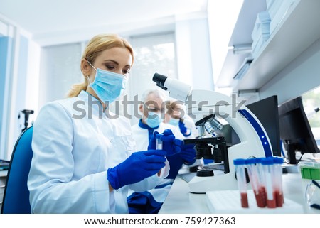 Scientific discovery. Blond young researcher sitting at the microscope and wearing a uniform and a medical face mask and holding test tubes while other researchers working in the background