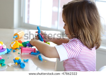 Little girl is learning to use colorful play dough in a well lit room near window