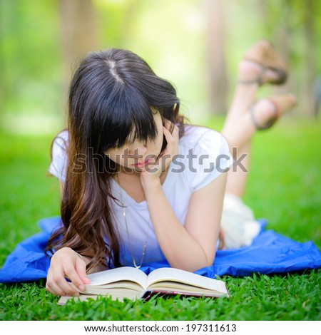 Beautiful happy Asian girl lies on meadow and reads the book , Happy smiling beautiful young university student studying lying down in grass.