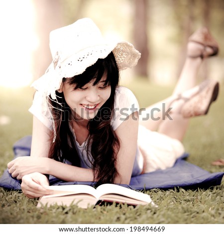 Beautiful happy Asian girl lies on meadow and reads the book , Happy smiling beautiful young university student studying lying down in grass. vintage portrait
