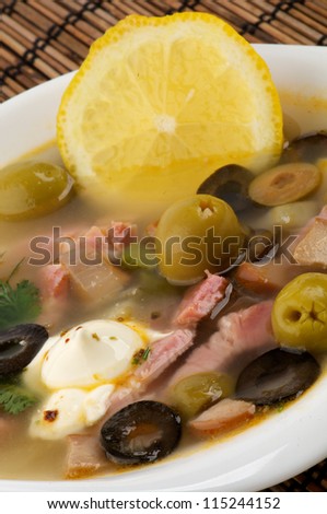 Russian Traditional Soup Solyanka with Lemon, Sour Cream and Green Olives in White Bowl closeup