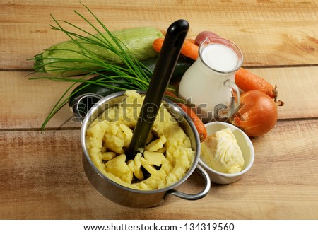 Preparing Vegetables Puree. Potato Masher in Potato, Jar of Milk, Butter, Onion, Carrot, Chive and Zucchini closeup on Wooden background