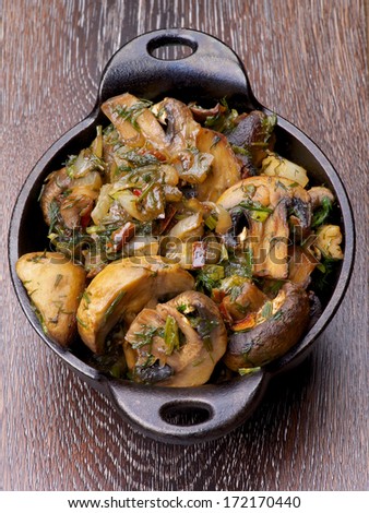 Delicious Champignon Mushrooms Stew with Onion and Greens in Black Cast-iron Stew Pot on Wooden background. Top View