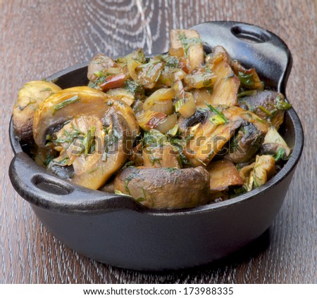Tasty Champignon Mushrooms Stew with Onion and Greens in Black Cast-iron Stew Pot on Wooden background