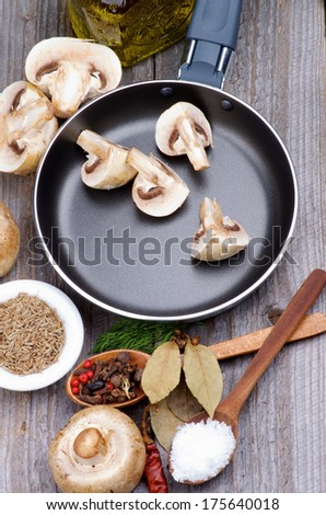 Preparing to Roast Edible Champignon Mushrooms in Frying Pan with Spices in Wooden Spoons  and Raw Ingredients closeup on Rustic Wooden background. Top View