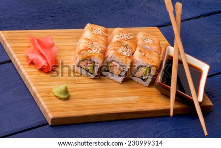 Delicious Salmon Maki Roll with Crab, Cucumber, Sesame Seeds and Soy Sauce, Ginger, Chop Sticks and Wasabi closeup on Straw Mat on Dark Blue Wooden background