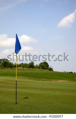 Golf green with hole, flagstick, bunker, blue sky, and wood in the background