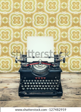 Retro Styled Image Of An Old Typewriter On A Wooden Floor With Vintage Wallpaper Behind It