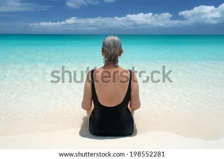 mature woman with gray hair sits at water\'s edge and looks out on ocean; back view with symmetry