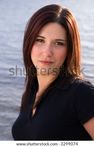 attractive young brunette woman head and shoulders shot, front view, against blue water. black polo shirt, serious expression