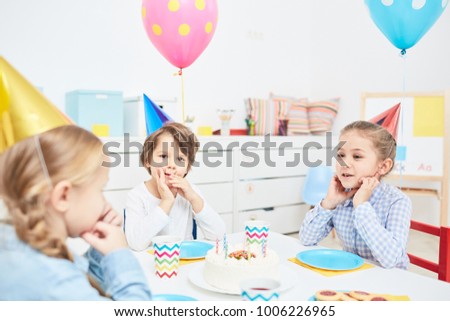 Cute little friends in birthday caps gathered by festive table with tasty cake in its center