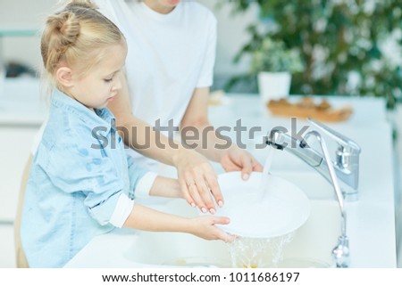 Little girl helping her mother wash dishes over sink in the kitchen after dinner