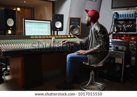 Young hipster sitting on chair in front of monitor and soundboard in studio of records