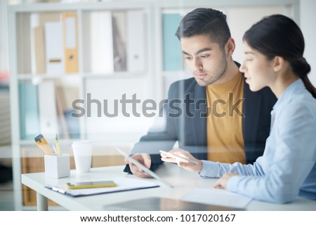 Young employee looking at touchpad display and listening to his colleague explanations of the data