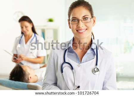 Portrait of a friendly physician smiling at camera, nurse and patient can be seen in background