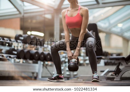 Fit young woman squatting while standing on the floor and holding heavy kettlebell between legs during training