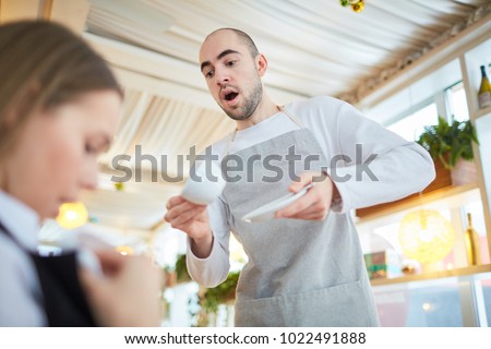 Careless waiter trying to catch cup of tea while young woman looking if there is stain on her clothes