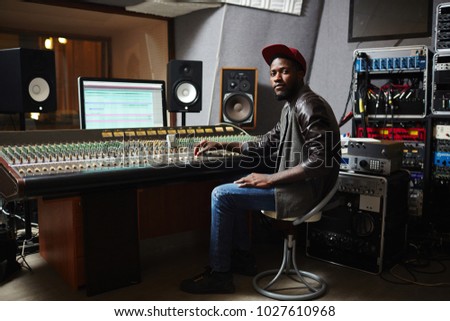 Young African-american rapper in baseball cap, jeans and leather jacket working by switchboard in studio